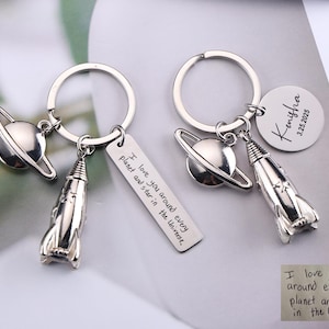Op de afbeelding: Twee zilveren sleutelhangers met ruimte-thema bedels. Elke sleutelhanger heeft een planeet, een raket en een label met de tekst "I love you around every planet and star in the Universe." Eén sleutelhanger heeft een rond label met een naam en datum.