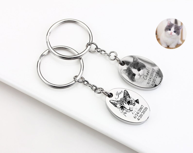 May include: Two silver keychains with oval pendants featuring cat portraits. One pendant has a grayscale photo of a kitten, the other a line drawing of a cat. Both pendants include dates and names. A small cat photo is in the upper right.