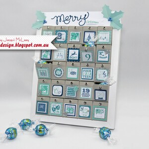 Puede incluir: Un calendario de adviento de madera blanca con 25 puertas numeradas. Cada puerta tiene un diseño diferente con un esquema de color azul y verde. El calendario dice "Feliz Navidad" en la parte superior. Hay varios chocolates envueltos en azul y plata delante del calendario.