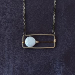 May include: A silver necklace with a rectangular frame and a white pearl pendant. The frame has a thin gold bar across the center.