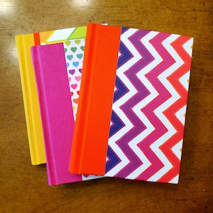 May include: A stack of colorful notebooks with hard covers. The notebooks feature a variety of designs, including a yellow cover, a pink cover, a cover with hearts, an orange cover, and a white cover with a zig-zag pattern in pink, purple, and red.