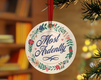 May include: A round, cream-colored Christmas ornament with a floral wreath design. The ornament features the text "Most Ardently" in blue script with a pink heart.