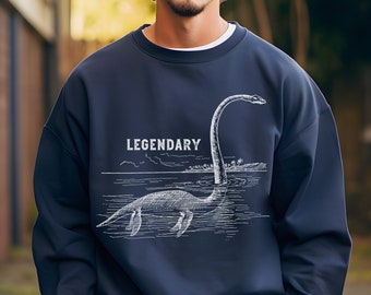 Puede incluir: Una sudadera azul marino con un gráfico blanco de un plesiosaurio nadando en el agua. El texto "LEGENDARY" está impreso sobre el plesiosaurio.