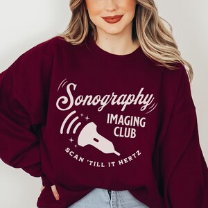 May include: A maroon sweatshirt with the words "Sonography Imaging Club" in white script. Below the text is a stylized sonogram image and the phrase "Scan 'till it Hertz". The sweatshirt is a crew neck style.