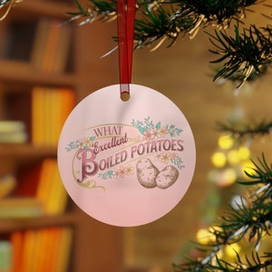 Könnte beinhalten: Ein runder, rosa Weihnachtsbaumschmuck mit dem Text "What Excellent Boiled Potatoes" in einer dekorativen Schriftart. Der Schmuck zeigt Illustrationen von Kartoffeln und floralen Akzenten, aufgehängt an einem roten Band.