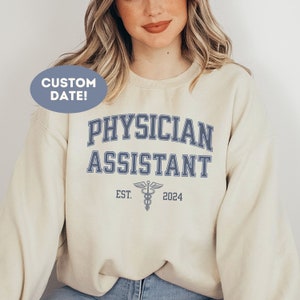 Può includere: Una felpa beige con la scritta "PHYSICIAN ASSISTANT" in blu con un simbolo di caduceo e la scritta "EST. 2024" sotto.