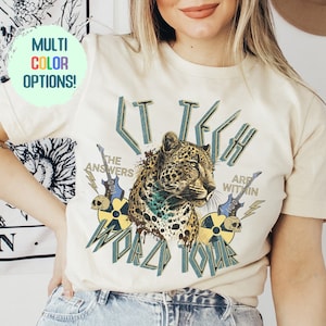 May include: A white t-shirt with a graphic print of a leopard, a skull, and a guitar. The text on the shirt reads "LT TECH WORLD TOUR" and "THE ANSWERS ARE WITHIN".
