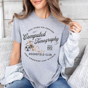 Puede incluir: Camiseta gris con un gr&aacute;fico de un esqueleto sosteniendo un carrete de pel&iacute;cula con el texto "You Pose We Expose Computed Tomography Est. 1972 The Hounsfield Club The Answers Are Within"