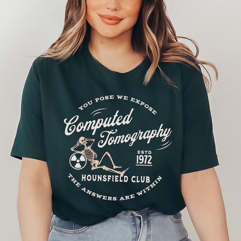 Puede incluir: Una camiseta verde oscuro con un dise&ntilde;o gr&aacute;fico blanco que presenta un esqueleto sentado en un c&iacute;rculo con el texto "Computed Tomography" y "The Hounsfield Club" en una fuente de estilo vintage.