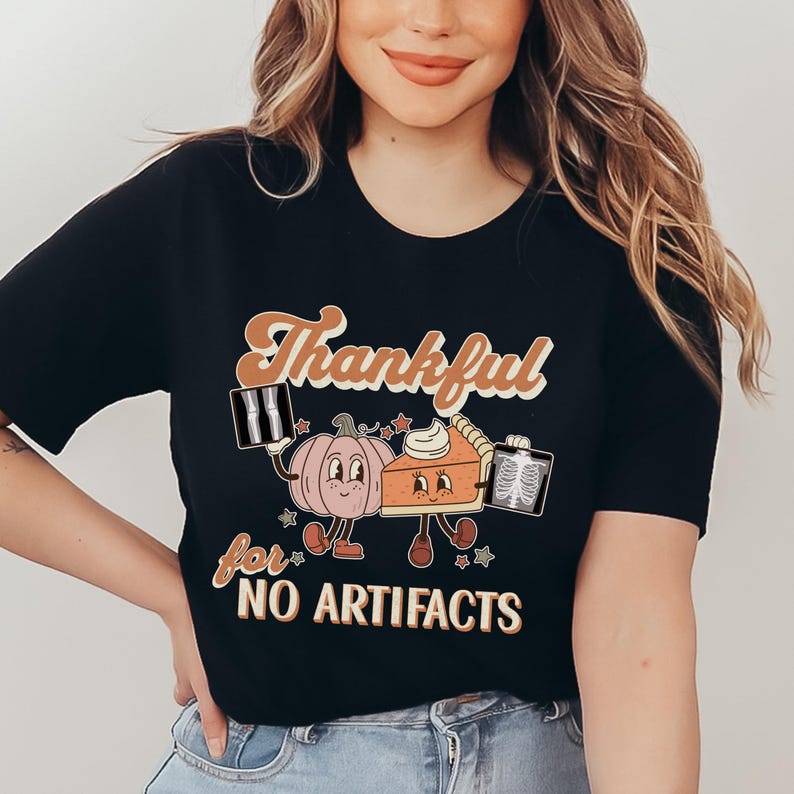 May include: Black t-shirt with a retro-style graphic of a pumpkin and a pie holding x-ray images. The text "Thankful for no artifacts" is printed below the graphic.