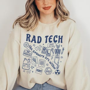 May include: Cream-colored sweatshirt with the words "RAD TECH" in navy blue, along with various medical-themed graphics. The design includes a skeleton, radiology symbols, and the phrase "the answers are within."