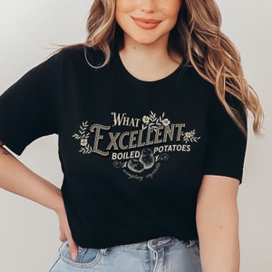 May include: Black t-shirt with a vintage-style design. The design features the words "What Excellent Boiled Potatoes" in a decorative font, along with illustrations of potatoes and floral accents. The phrase "exemplary vegetable" is also included.