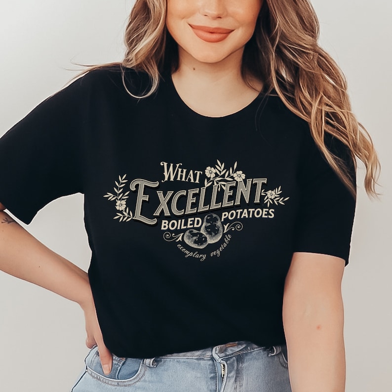 May include: Black t-shirt with a vintage-style design. The design features the words "What Excellent Boiled Potatoes" in a decorative font, along with illustrations of potatoes and floral accents. The phrase "exemplary vegetable" is also included.