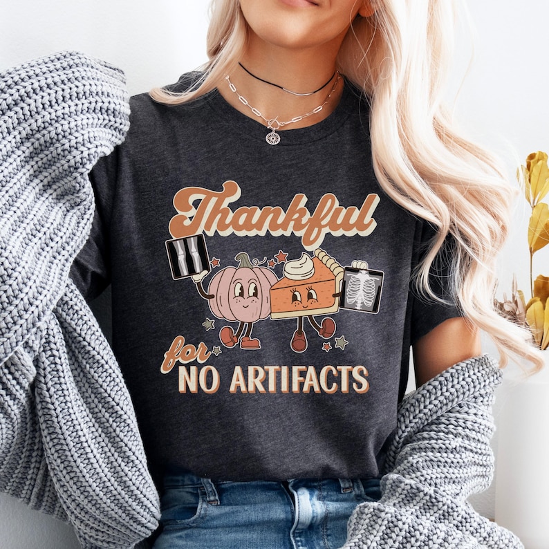 May include: A dark gray t-shirt with a retro-style graphic of a pumpkin and a pie holding x-ray images. The text on the shirt reads "Thankful for no artifacts".