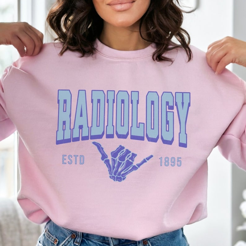 May include: Pink sweatshirt with the text "RADIOLOGY ESTD 1895" in blue and purple lettering. A skeleton hand with a hang loose gesture is in the center of the text.