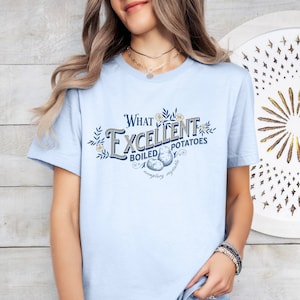 May include: Light blue t-shirt with the text "What Excellent Boiled Potatoes" in a vintage-style design. The graphic includes illustrations of potatoes and floral accents. The shirt is a short-sleeved crew neck.