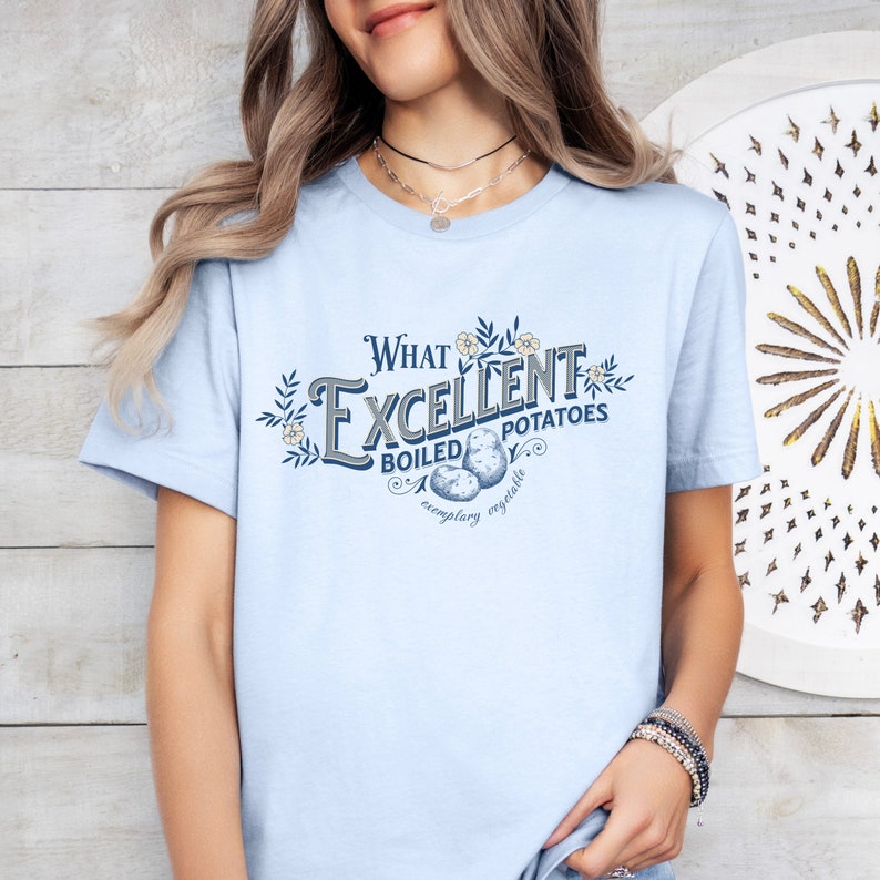 May include: Light blue t-shirt with the text "What Excellent Boiled Potatoes" in a vintage-style design. The graphic includes illustrations of potatoes and floral accents. The shirt is a short-sleeved crew neck.