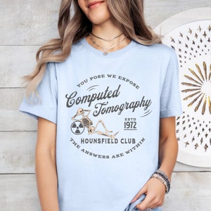 Puede incluir: Camiseta azul claro con un gr&aacute;fico de un esqueleto sentado en una rueda con un s&iacute;mbolo de radiaci&oacute;n. El texto dice "You Pose We Expose Computed Tomography Estd 1972 The Hounsfield Club The Answers Are Within"