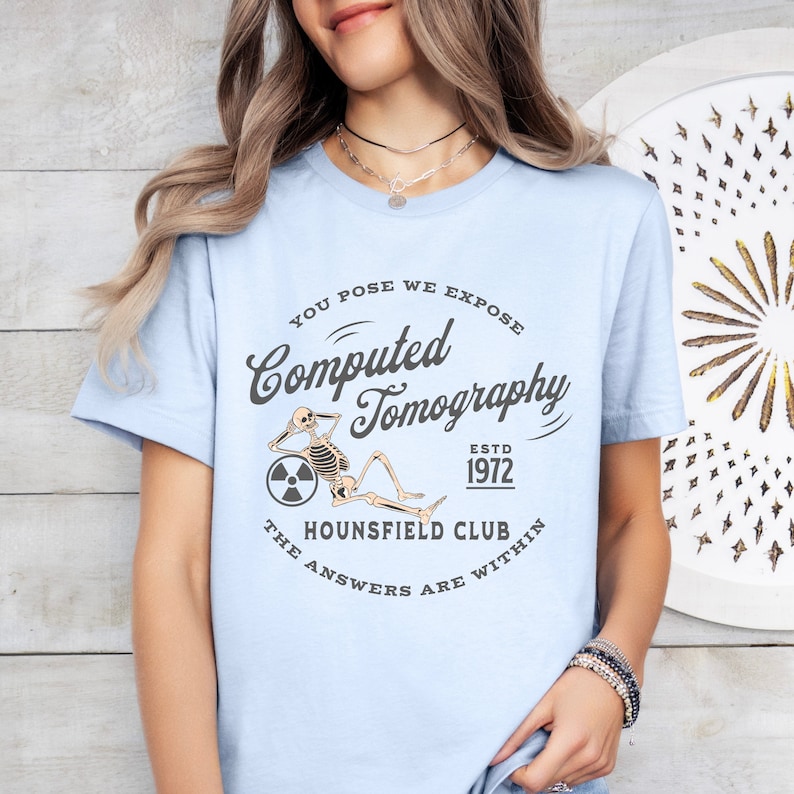 Puede incluir: Camiseta azul claro con un gr&aacute;fico de un esqueleto sentado en una rueda con un s&iacute;mbolo de radiaci&oacute;n. El texto dice "You Pose We Expose Computed Tomography Estd 1972 The Hounsfield Club The Answers Are Within"