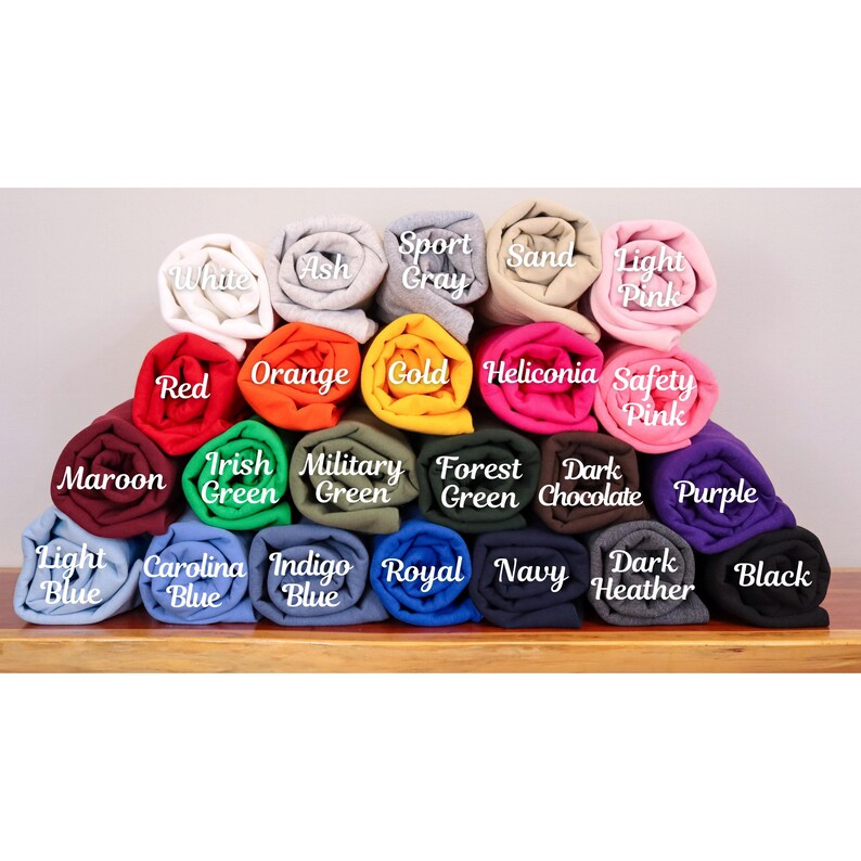 May include: A display of rolled-up fabric in various colors, including white, red, orange, gold, and purple. Each roll is labeled with its color name, such as "Navy," "Black," and "Light Pink." The fabric is stacked on a wooden surface.