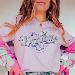 May include: A light purple t-shirt with a graphic design that says "What Excellent Boiled Potatoes, exemplary vegetable". The graphic features a vintage style font and illustrations of potatoes.