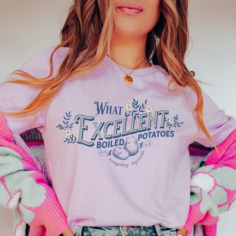 May include: A light purple t-shirt with a graphic design that says "What Excellent Boiled Potatoes, exemplary vegetable". The graphic features a vintage style font and illustrations of potatoes.