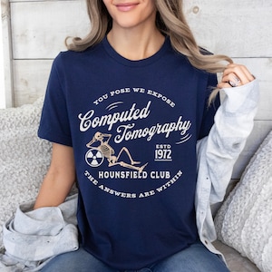 Puede incluir: Una camiseta azul marino con un gr&aacute;fico blanco de un esqueleto sentado en una rueda con un s&iacute;mbolo de radiaci&oacute;n. El texto en la camiseta dice "You Pose We Expose Computed Tomography Estd 1972 Hounsfield Club The Answers Are Within".