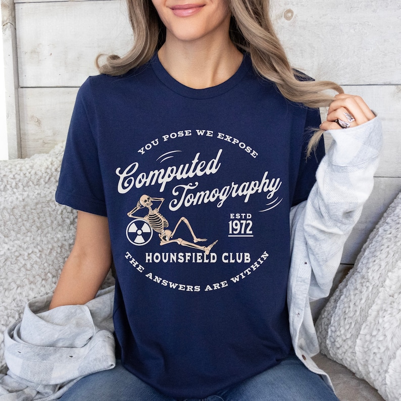 Puede incluir: Una camiseta azul marino con un gr&aacute;fico blanco de un esqueleto sentado en una rueda con un s&iacute;mbolo de radiaci&oacute;n. El texto en la camiseta dice "You Pose We Expose Computed Tomography Estd 1972 Hounsfield Club The Answers Are Within".