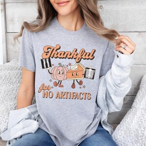 May include: Gray t-shirt with a retro-style graphic of a pumpkin and a pie holding x-rays. The text reads "Thankful for NO ARTIFACTS".