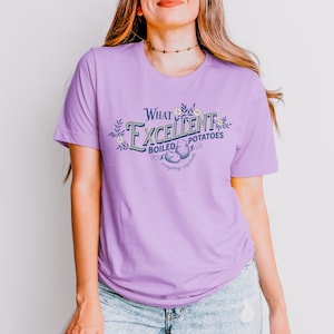 May include: A light purple t-shirt with the text "What Excellent Boiled Potatoes" in a vintage-style design. The graphic includes illustrations of potatoes and floral accents. The shirt is made of a soft material.
