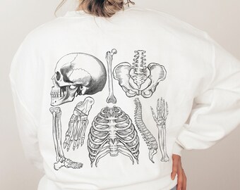 Puede incluir: Una sudadera blanca con una ilustración anatómica en blanco y negro de un esqueleto humano en la espalda. La ilustración incluye un cráneo, una caja torácica, una columna vertebral, una pelvis y huesos de la mano.