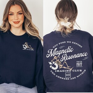 May include: A navy blue sweatshirt with a white graphic on the front and back. The front graphic is a skeleton sitting on a magnet with the text "AMY RT(R)(MR)". The back graphic is a skeleton sitting on a magnet with the text "YOU POSE WE EXPOSE Magnetic Resonance ESTD 1977 THE IMAGING CLUB MR ANSWERS ARE WITHIN".