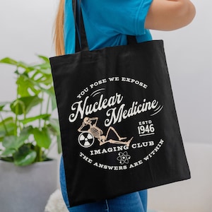 May include: Black tote bag with a white graphic design featuring a skeleton holding a radiation symbol and the text "You pose we expose Nuclear Medicine Est. 1946 Imaging Club The answers are within".