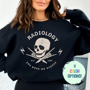 May include: Black sweatshirt with a white skull and crossbones graphic. The graphic says "Radiology" with the text "EST 1895 You Pose We Expose".