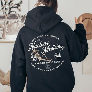 Puede incluir: Sudadera con capucha negra con un estampado gráfico blanco en la espalda. El gráfico presenta un esqueleto sentado en una rueda con el texto "You Pose We Expose Nuclear Medicine Est. 1946 The Imaging Club The Answers Are Within".