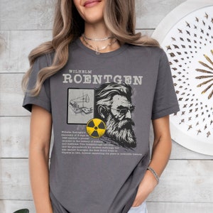 Op de afbeelding: Een grijs T-shirt met een zwart-witte illustratie van Wilhelm Conrad Röntgen, een Duitse natuurkundige, en een citaat over zijn ontdekking van röntgenstralen in 1895. Het shirt bevat ook een geel en zwart stralingssymbool.