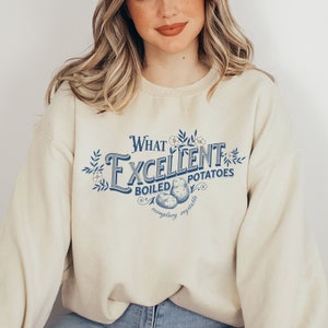 Puede incluir: Una sudadera beige con un estampado gráfico azul que dice "What Excellent Boiled Potatoes" con un dibujo de patatas y flores.