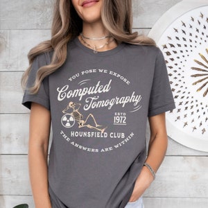 Puede incluir: Una camiseta gris oscuro con un dise&ntilde;o gr&aacute;fico blanco que presenta un esqueleto sentado en un c&iacute;rculo con el texto "You Pose We Expose Computed Tomography Est. 1972 The Hounsfield Club The Answers Are Within"