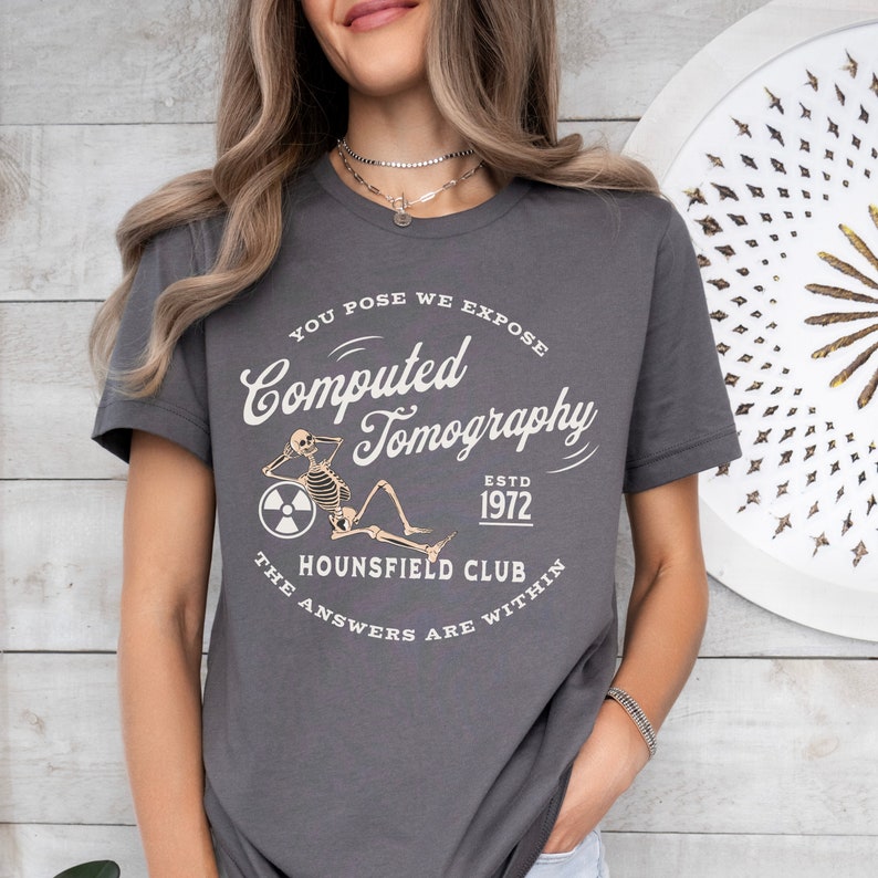 Puede incluir: Una camiseta gris oscuro con un dise&ntilde;o gr&aacute;fico blanco que presenta un esqueleto sentado en un c&iacute;rculo con el texto "You Pose We Expose Computed Tomography Est. 1972 The Hounsfield Club The Answers Are Within"