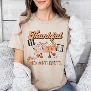 May include: A beige t-shirt with a graphic of a pumpkin and a pie holding x-rays. The text on the shirt reads "Thankful for NO ARTIFACTS".