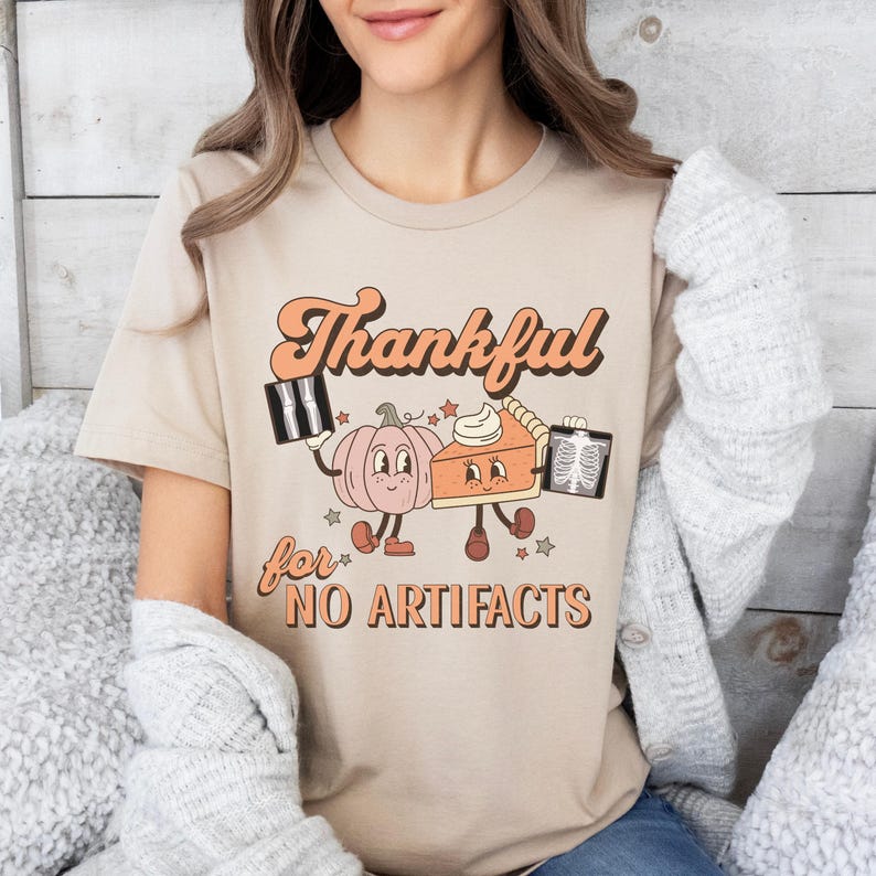 May include: A beige t-shirt with a graphic of a pumpkin and a pie holding x-rays. The text on the shirt reads "Thankful for NO ARTIFACTS".