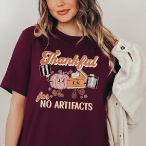 May include: A maroon t-shirt with a graphic of a pumpkin and a pie holding x-rays. The text on the shirt reads "Thankful for NO ARTIFACTS".