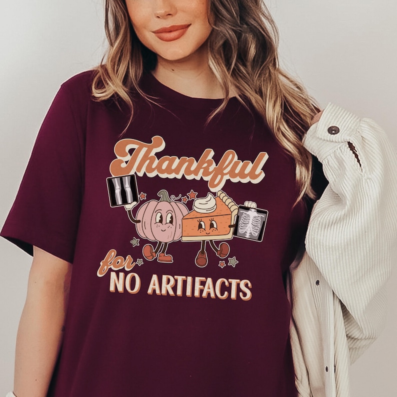 May include: A maroon t-shirt with a graphic of a pumpkin and a pie holding x-rays. The text on the shirt reads "Thankful for NO ARTIFACTS".