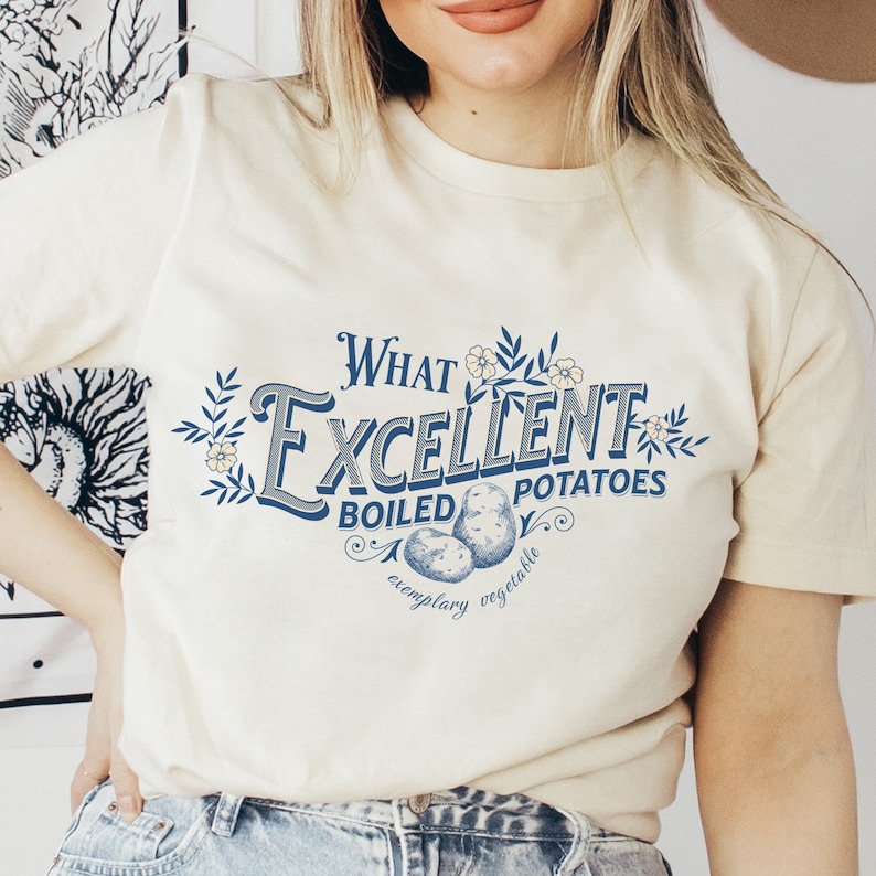 May include: Cream-colored t-shirt featuring the text "What Excellent Boiled Potatoes" in a vintage-style design with blue and white accents. The graphic includes illustrations of potatoes and floral elements.