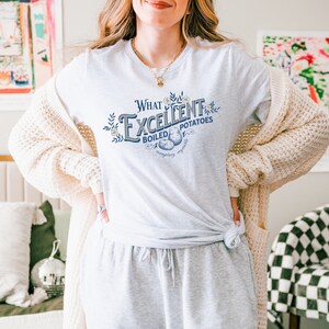 May include: A light gray t-shirt with the text "What Excellent Boiled Potatoes" in a decorative font. The shirt features a graphic of potatoes and floral accents. The person is wearing gray sweatpants and a cream-colored cardigan.