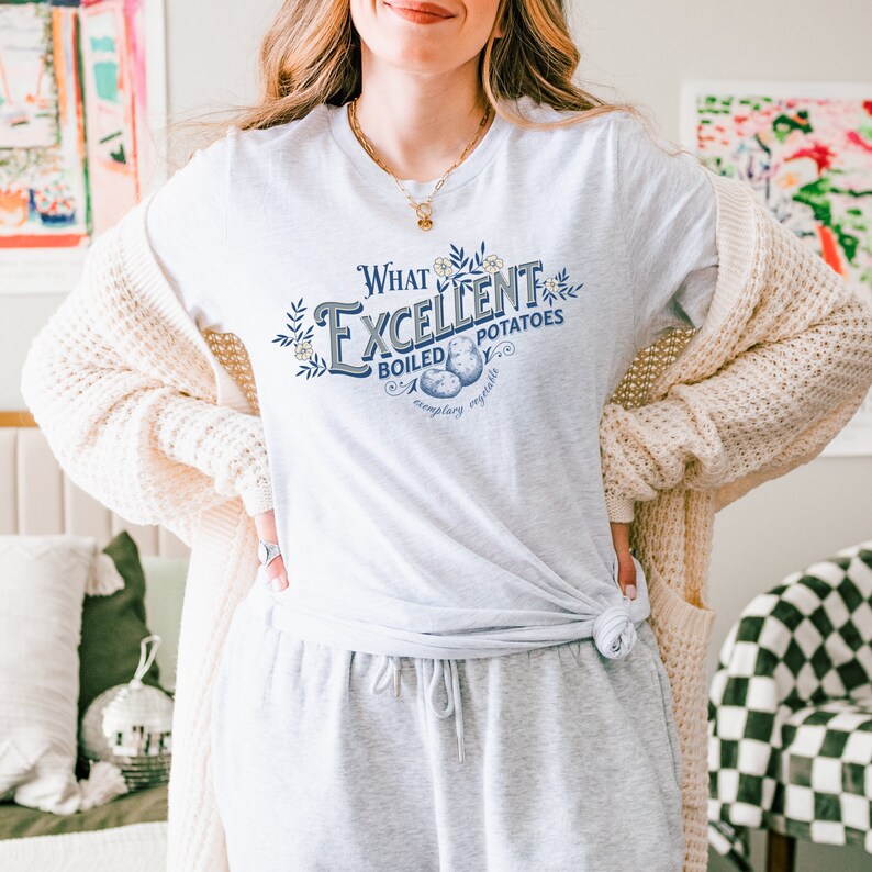May include: A light gray t-shirt with the text "What Excellent Boiled Potatoes" in a decorative font. The shirt features a graphic of potatoes and floral accents. The person is wearing gray sweatpants and a cream-colored cardigan.