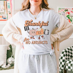 May include: A gray t-shirt with a retro design featuring a pumpkin and a pie with the text "Thankful for no artifacts".