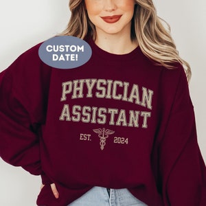 Può includere: Felpa color borgogna con il testo "PHYSICIAN ASSISTANT" in lettere dorate, un simbolo di caduceo e il testo "EST. 2024".