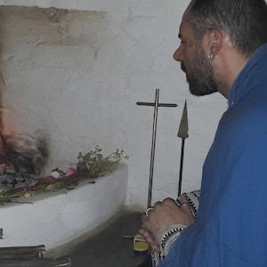May include: A person wearing a blue cloak with patterned sleeves sits before a stone fireplace with a burning fire. On the hearth, there are scattered flowers, greenery, a metal cross, and a spear-like object against a white wall.