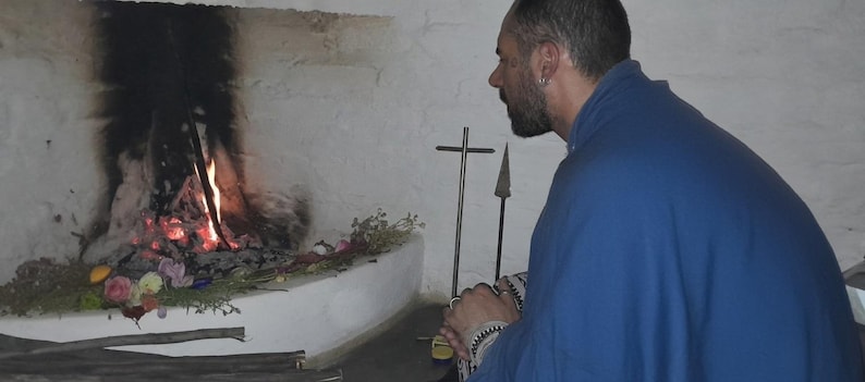 May include: A person wearing a blue cloak with patterned sleeves sits before a stone fireplace with a burning fire. On the hearth, there are scattered flowers, greenery, a metal cross, and a spear-like object against a white wall.